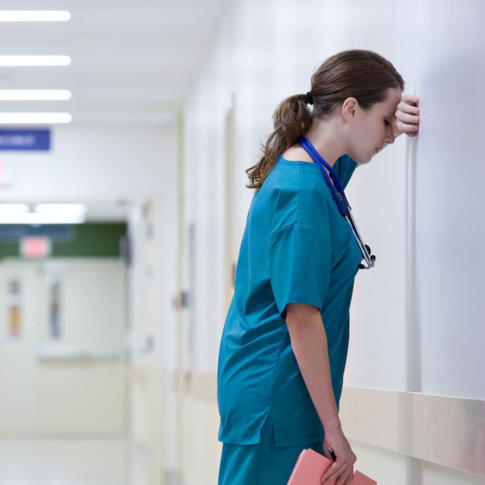 Foto: Krankenschwester lehnt mit dem Gesicht zu einer Wand. Mit einer Hand stützt sie sich ab.