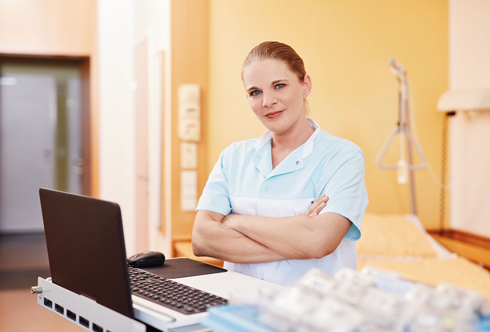 Foto: Krankenschwester steht mit verschränkten Armen vor einem Laptop in einem Krankenzimmer.